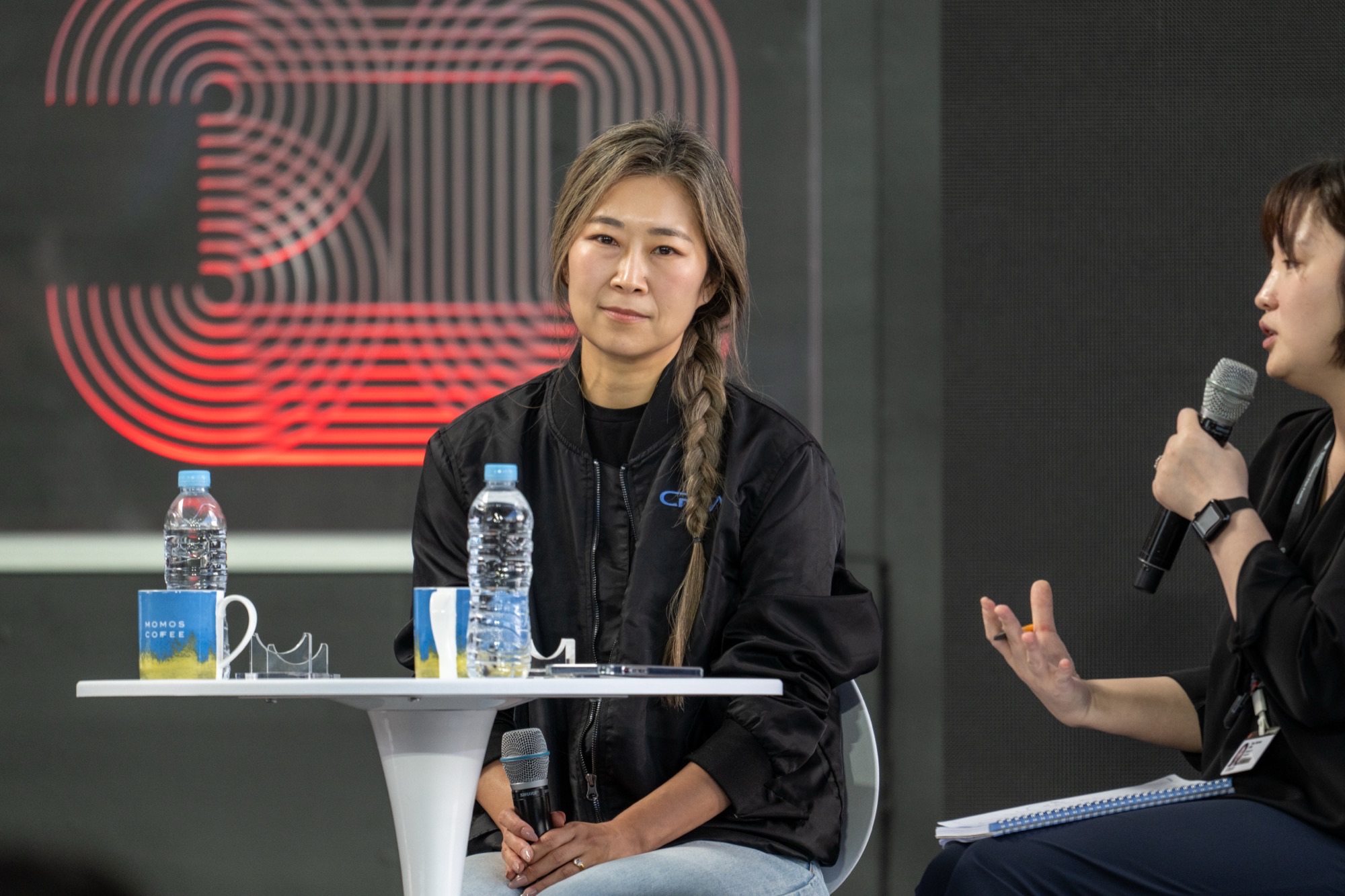 Maggie Kang sits on stage with a microphone during a panel appearance, wearing a black jacket with her hair in a braid.