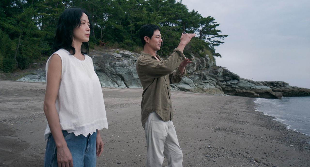 Jeon Do-yeon in a white sleeveless top stands on a beach beside Zo In-sung, who gestures toward the trees, with a rocky cliff and the sea behind them.