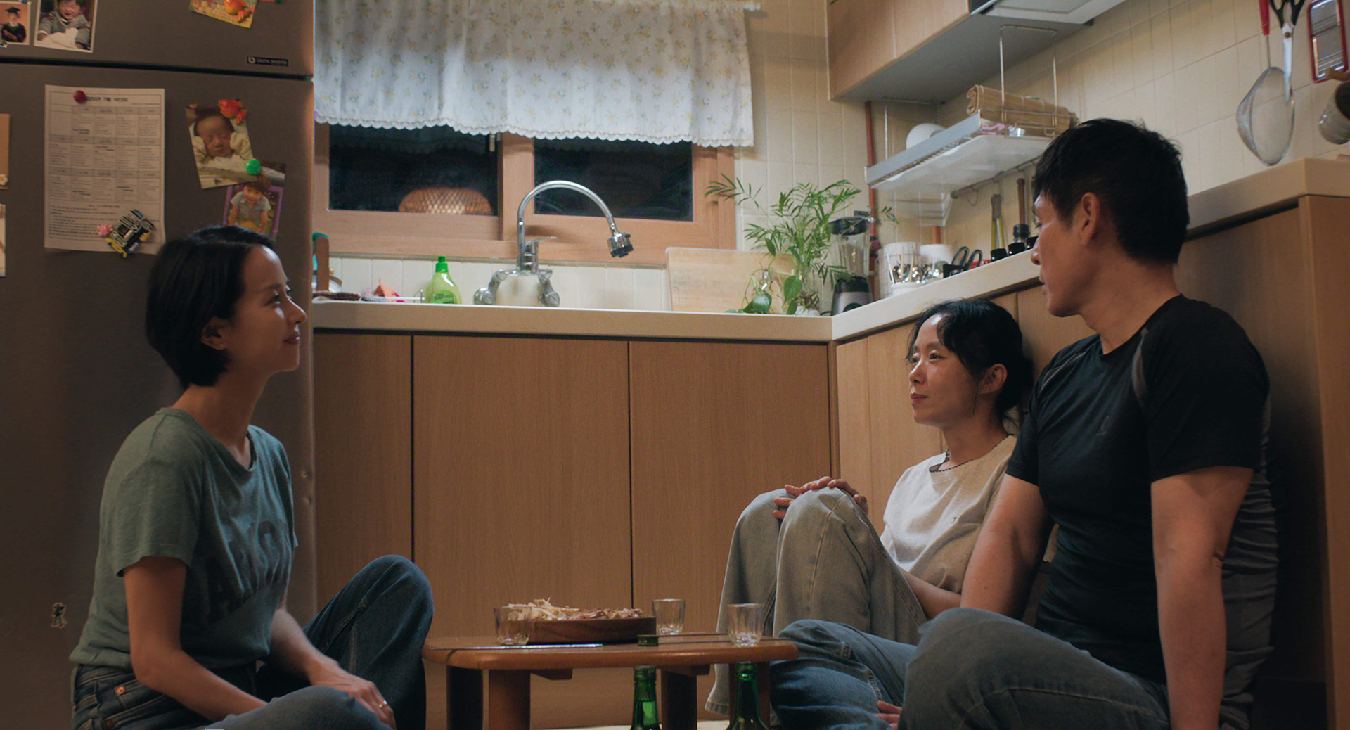 Jeon Do-yeon as Mi-ok sits on a kitchen floor between Cho Yeo-jeong and Sul Kyung-gu, with soju bottles and a low table in front of them.