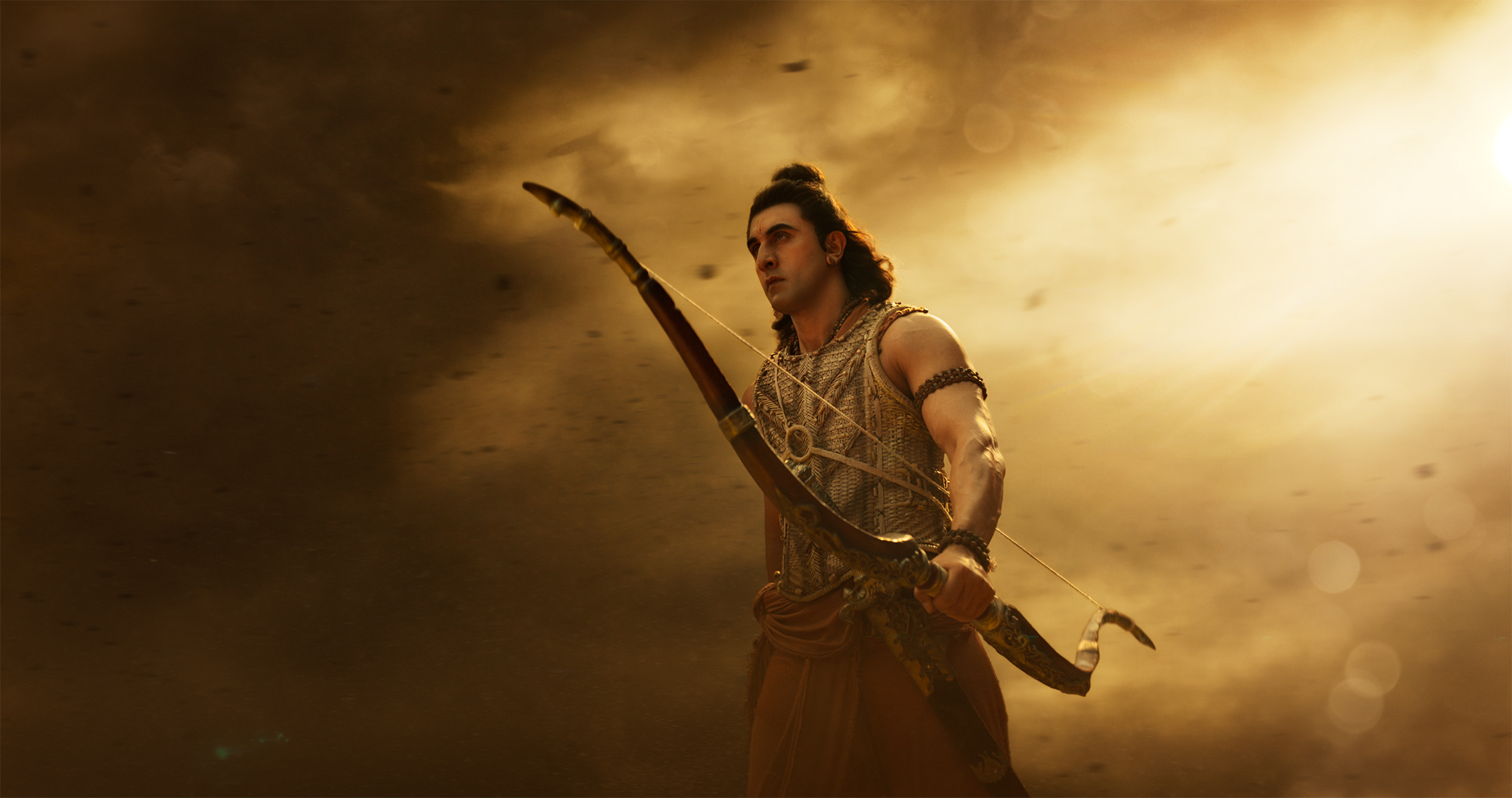 Ranbir Kapoor as Rama in Ramayana, hair tied up, holding a bow in gold light and dust.