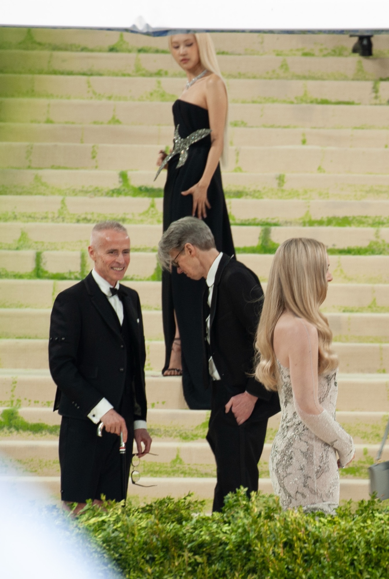 Rosé stands on the Metropolitan Museum steps at the 2026 Met Gala in a black strapless gown with a jeweled waist, guests in black tie in the foreground.