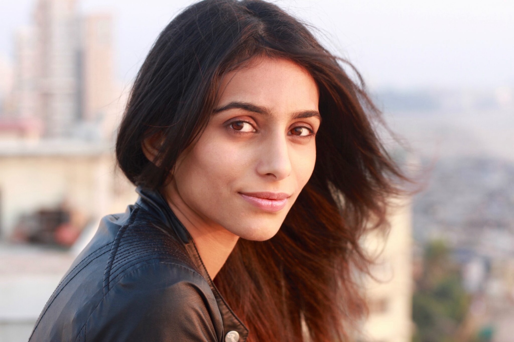 Shreya Patel in a black leather jacket, photographed outdoors against a soft city skyline at golden hour.