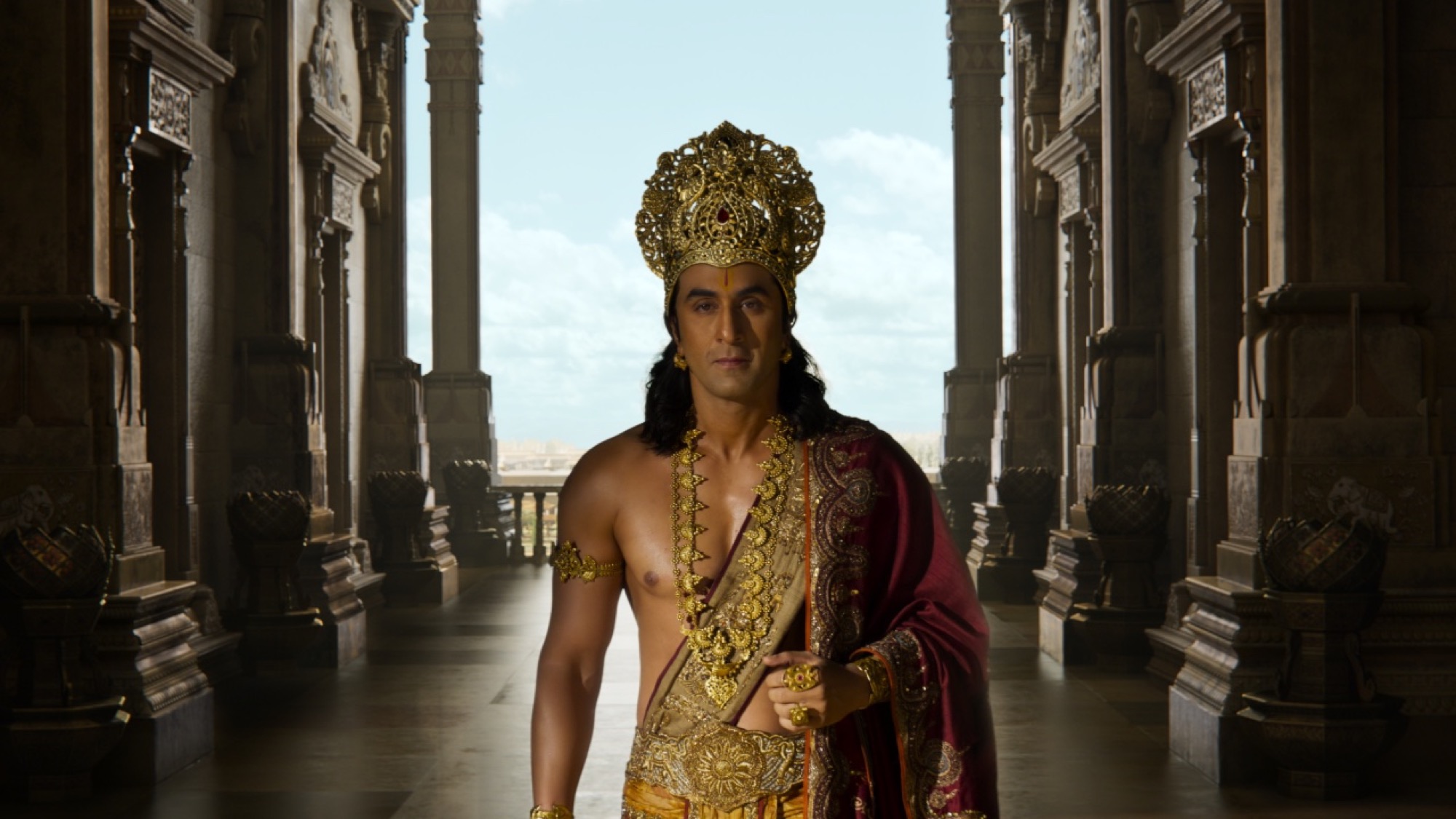 Ranbir Kapoor as Rama in gold crown and jewelry, standing in a carved palace corridor for Prime Focus Studios' Ramayana.