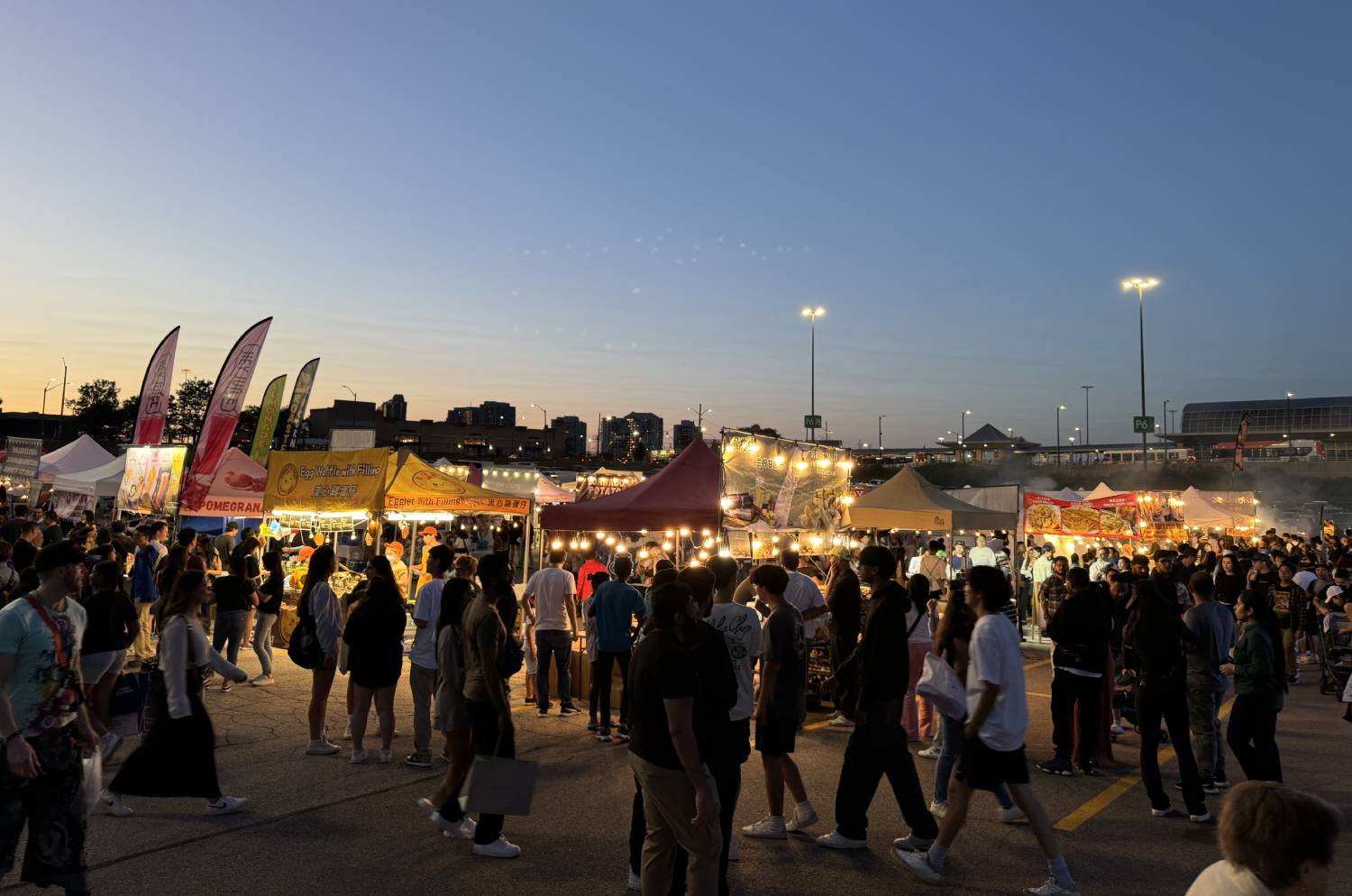 Crowds walking between Asian street-food tents under string lights at dusk at an outdoor night market.