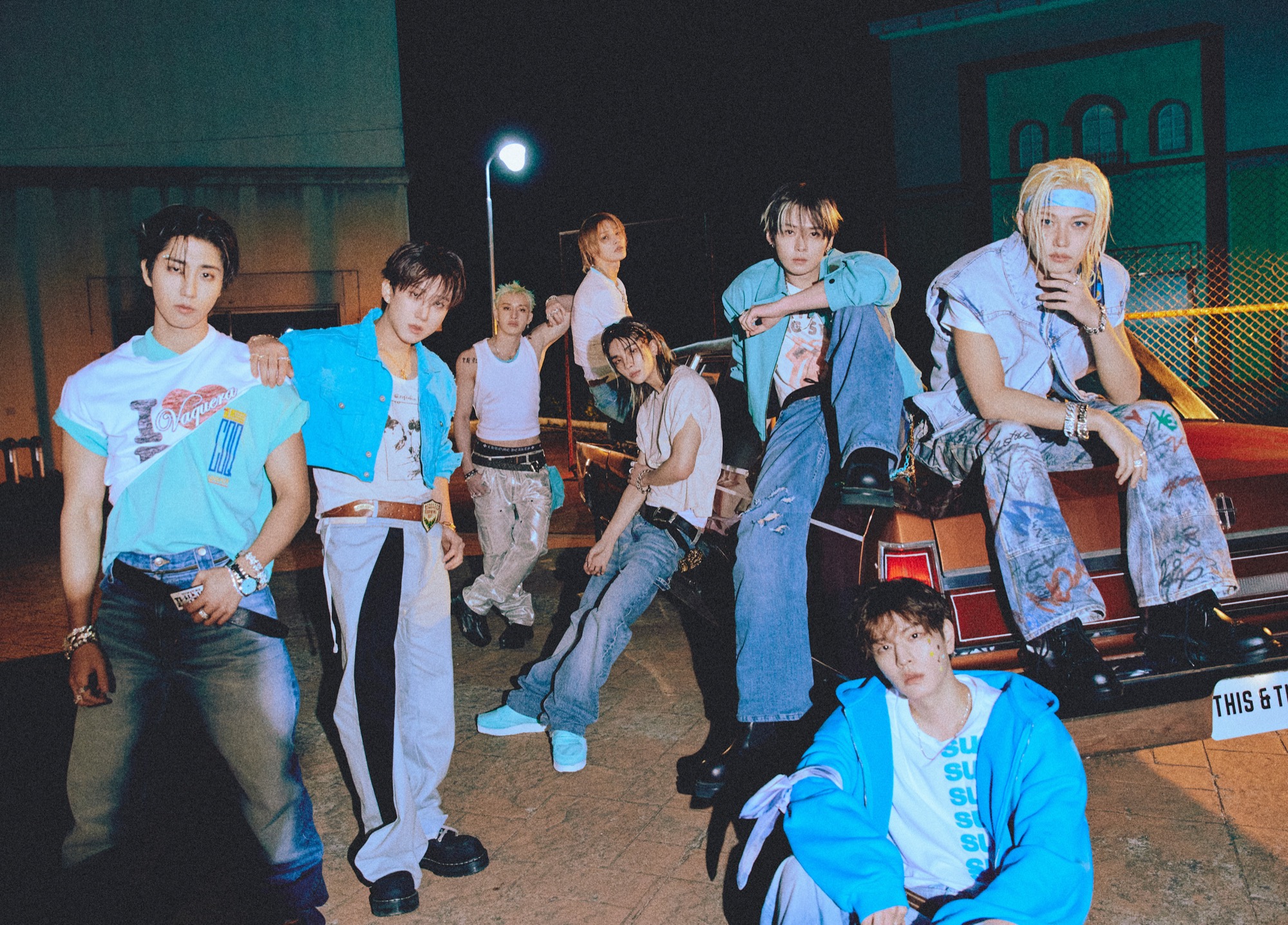 The eight members of Stray Kids gather around a red car in blue and white outfits for the THIS AND THAT album campaign.
