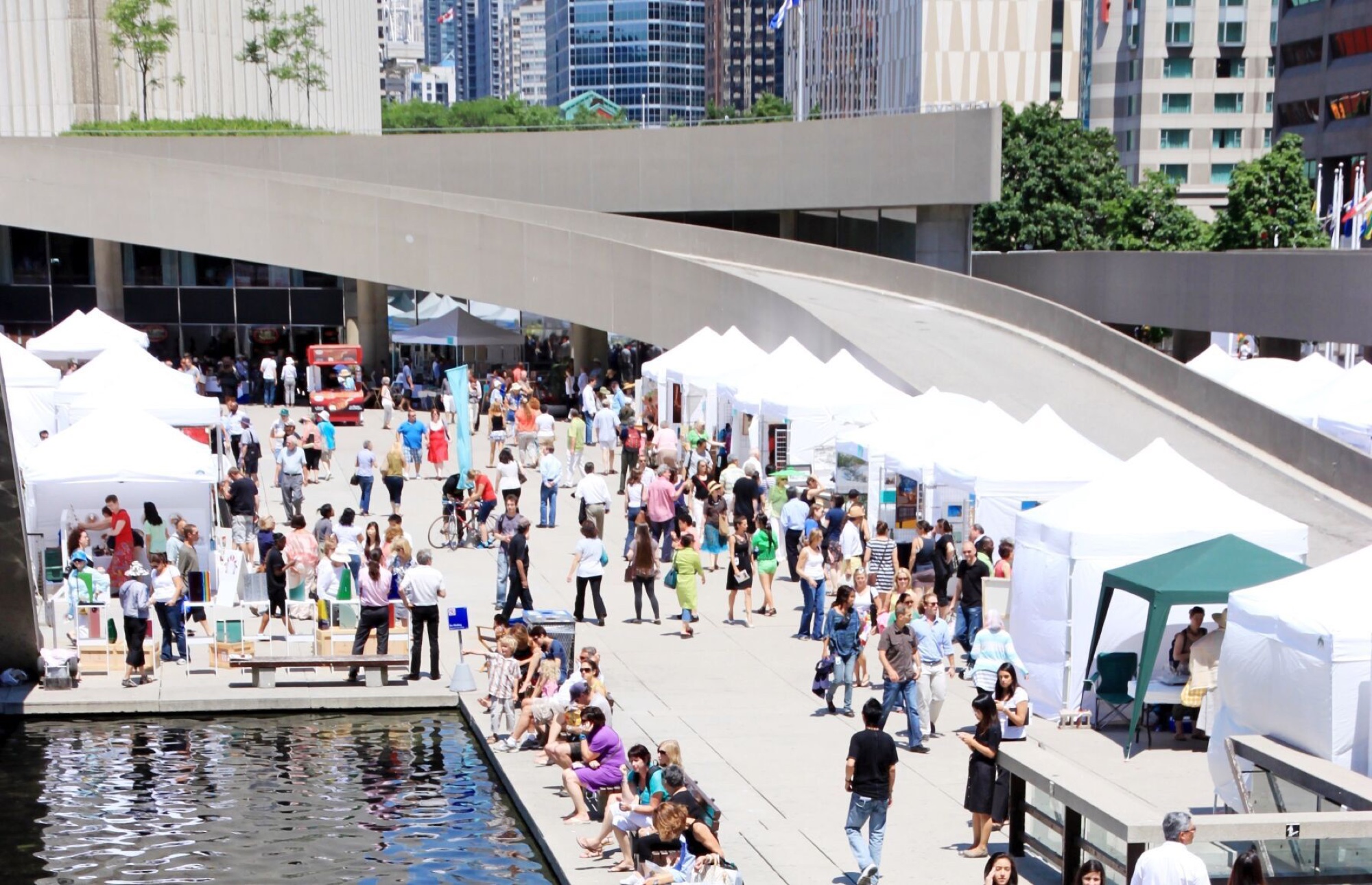 Crowds and white vendor tents filling Nathan Phillips Square under the curved concrete ramp in downtown Toronto.