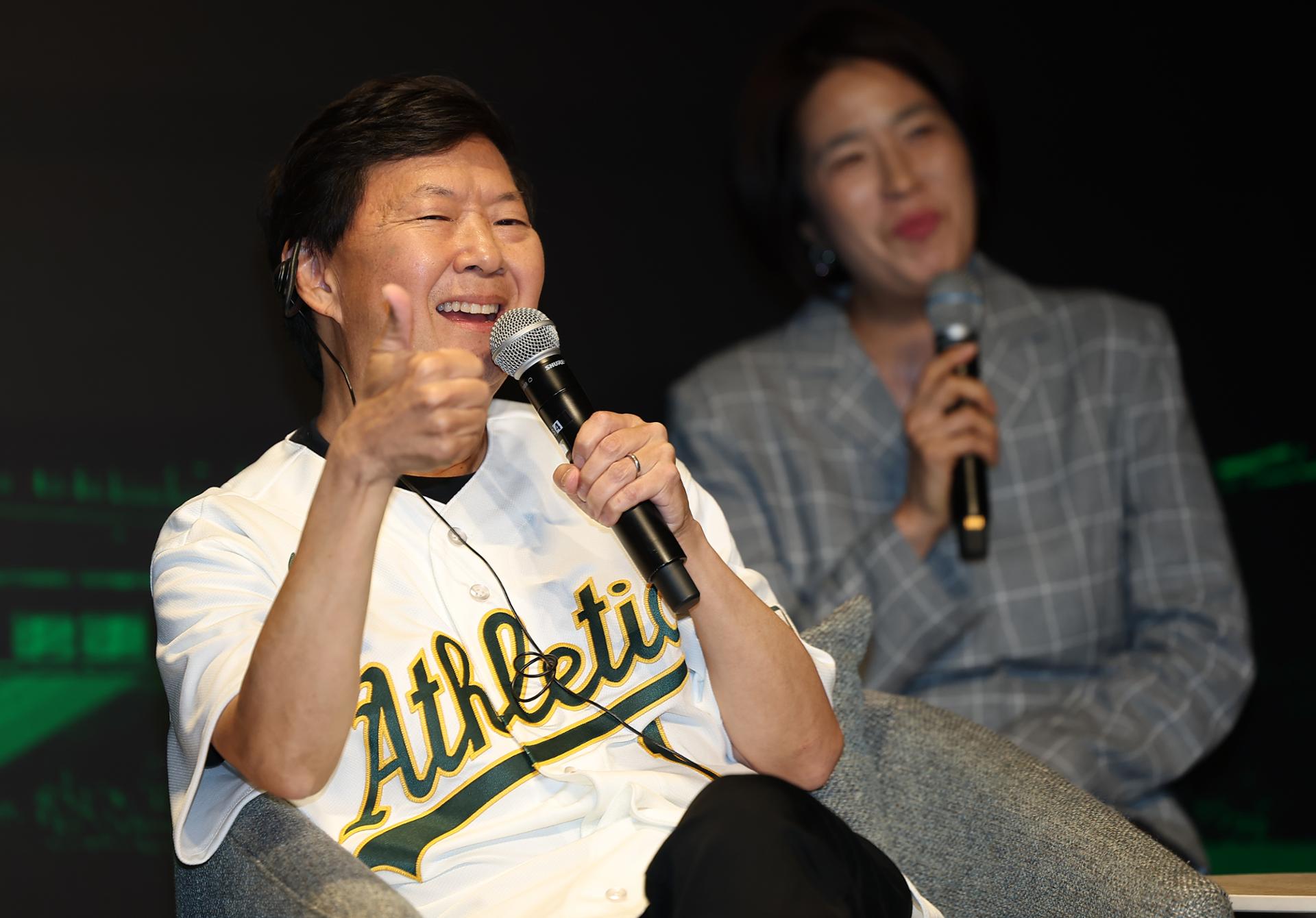 Ken Jeong in an Athletics jersey, smiling and giving a thumbs-up with a microphone at the Team61 press conference in Seoul.