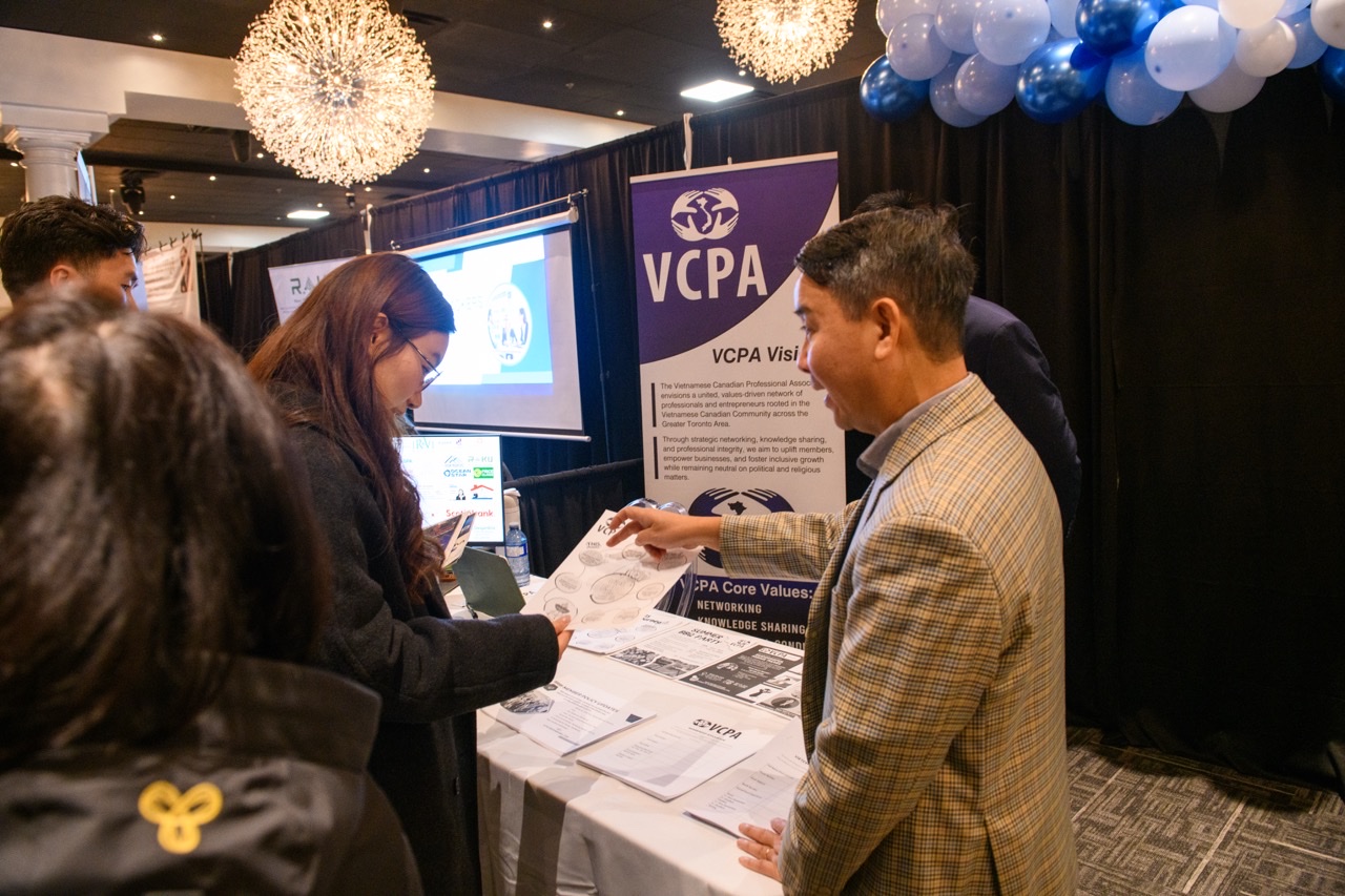 VCPA booth at Viet Expo, with a purple association banner, balloons, and visitors talking over flyers under Oasis-style chandeliers.