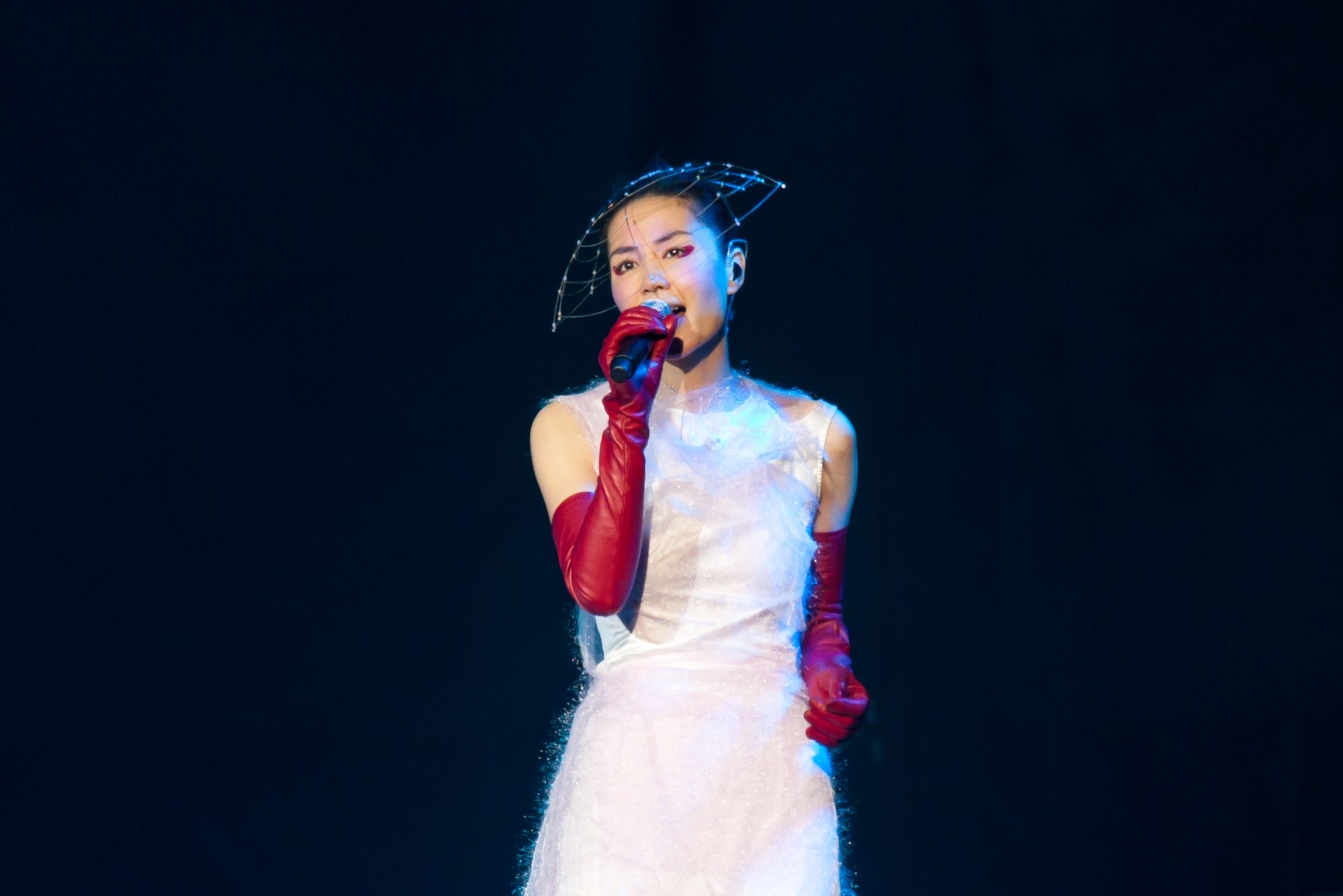 Faye Wong singing into a microphone on a dark stage, in a white dress, long red gloves, and a wire headpiece.