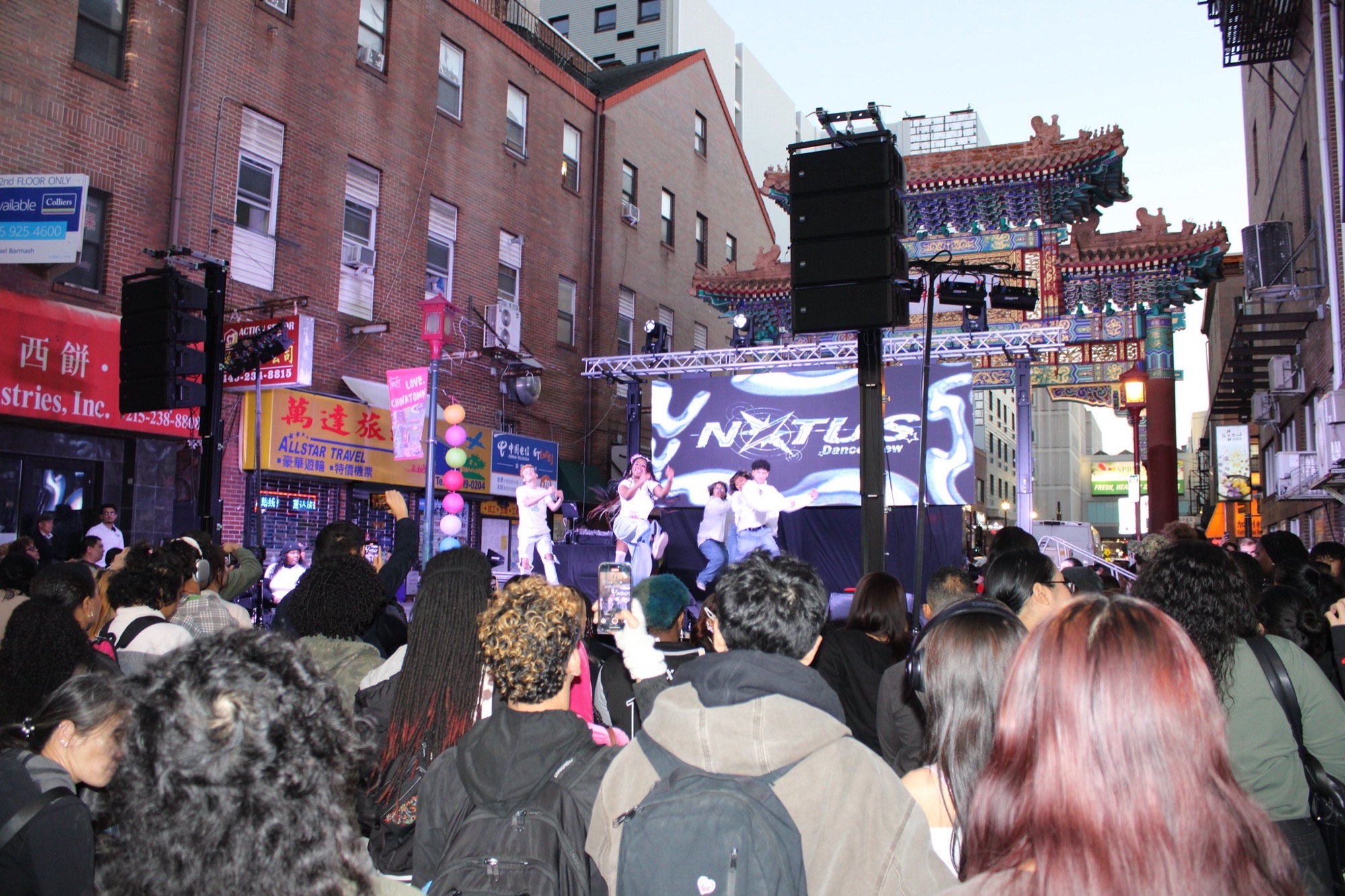 NXTUS Dance Crew performs on an outdoor stage near Philadelphia Chinatown's Friendship Gate during a prior YèShì Night Market, with lanterns and a street crowd in front of the brick storefronts.