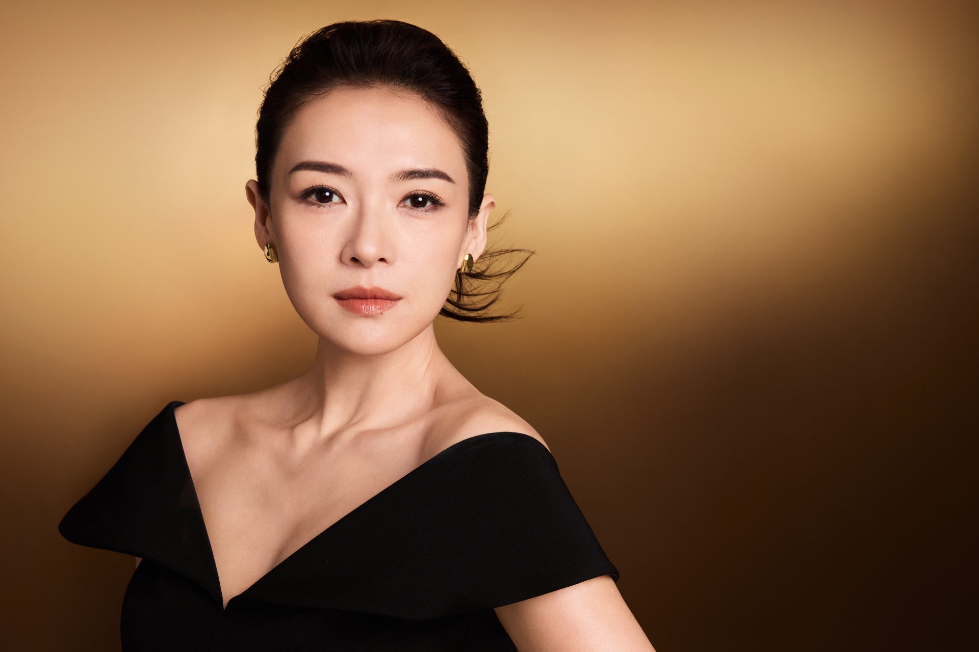 Zhang Ziyi in a black off-shoulder dress against a warm gold backdrop for Clarins Precious.